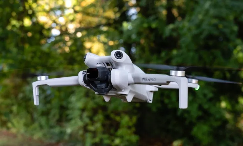Close-up of a white DJI Mini 4 Pro drone in flight against a blurred green foliage background.