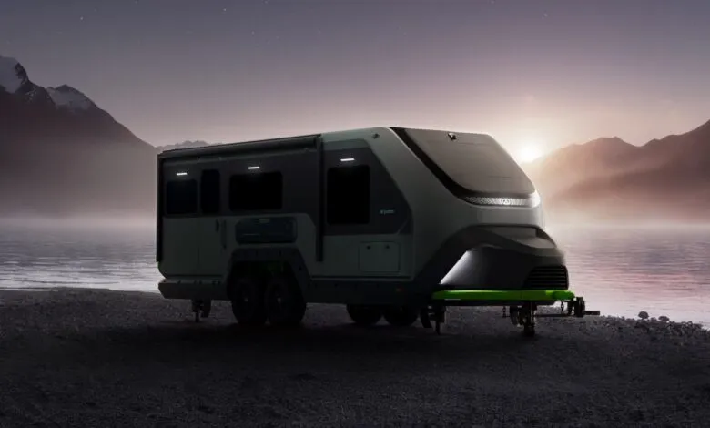 Futuristic RV trailer parked by a calm lake at dusk with mountains in the background.