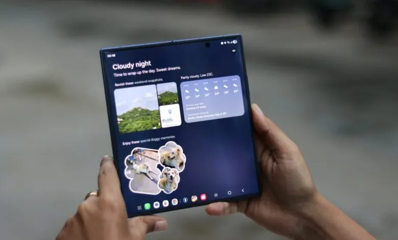 Hands holding a large, unfolded smartphone displaying a dark blue interface with weather and photo widgets.