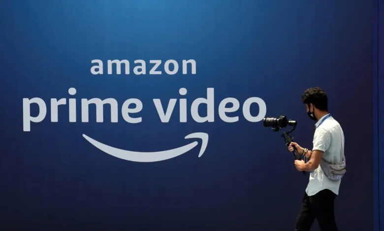 A person with a camera rig walks past a large Amazon Prime Video logo on a blue wall.