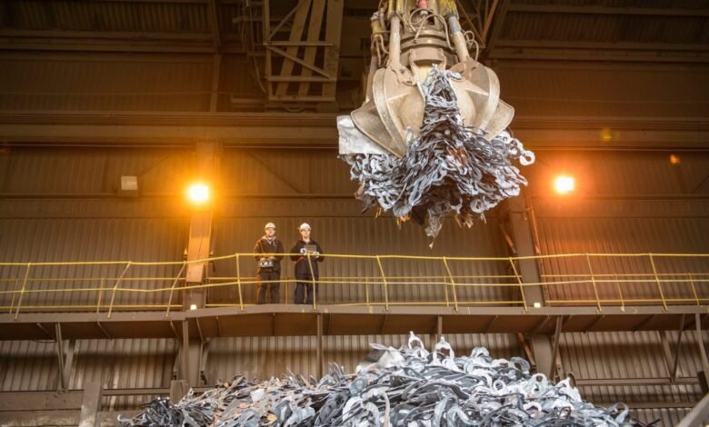 Two workers in hard hats observe a large industrial claw lifting scrap metal in a factory.