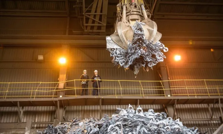 Two workers in hard hats observe a large industrial claw lifting scrap metal in a factory.