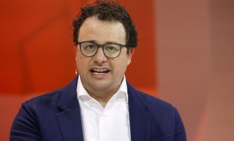 Man with curly hair and glasses speaking, wearing a blue blazer over a white shirt.