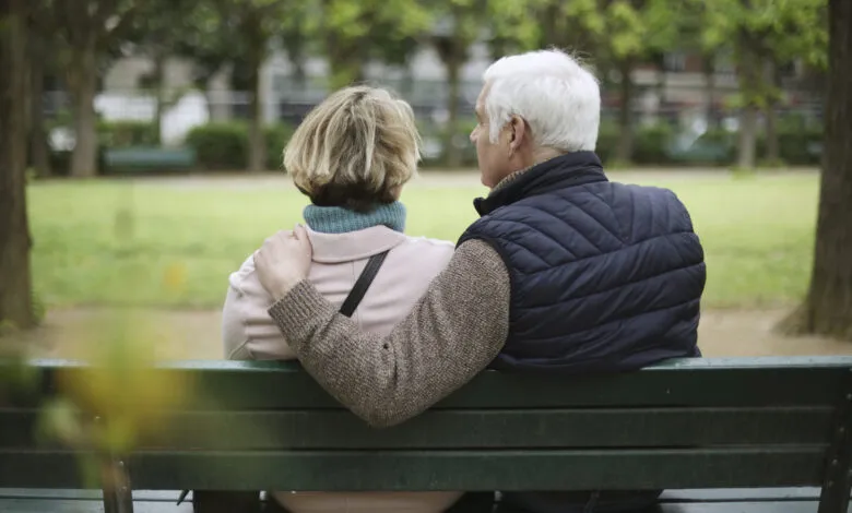 Elderly couple embracing on a park bench, with the man's arm around the woman.