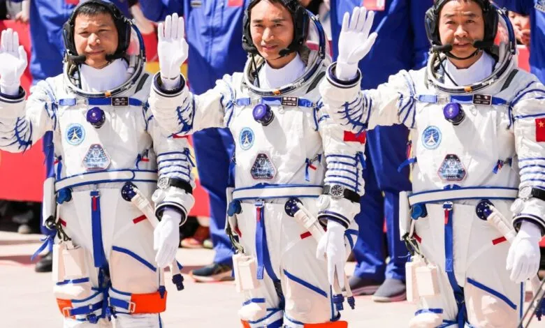 Three Chinese astronauts in white and blue spacesuits wave with both hands raised.