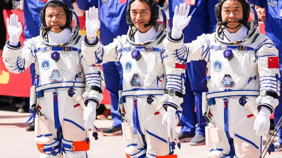 Three Chinese astronauts in white and blue spacesuits wave with both hands raised.
