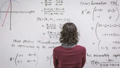 A person with curly hair looks at complex mathematical equations written on a whiteboard.