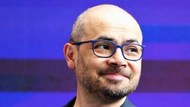 Close-up of a bald man with a beard wearing blue-rimmed glasses, looking to the side.