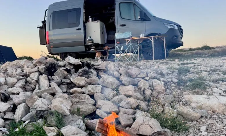 A beagle dog stands near a camper van with an open door, a campfire burning in the foreground.