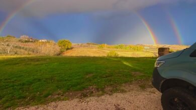 A double rainbow arches over rolling hills with a teal van in the foreground.