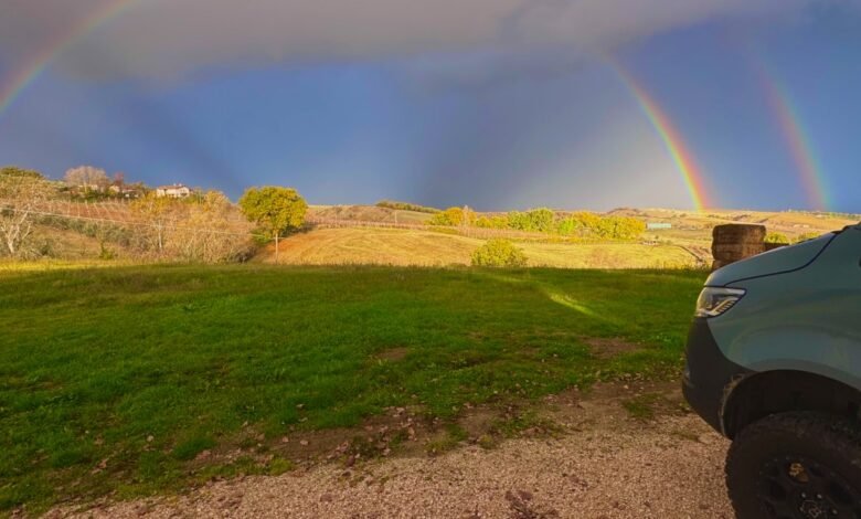 A double rainbow arches over rolling hills with a teal van in the foreground.