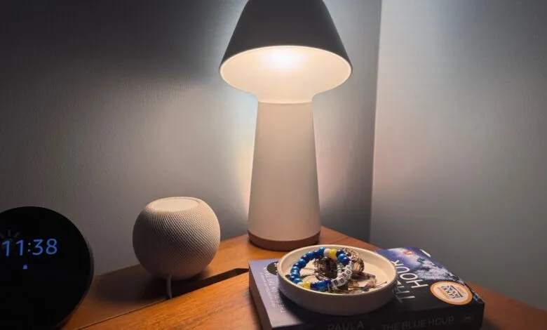 A bedside table with a smart speaker, alarm clock, lamp, and a dish of jewelry.