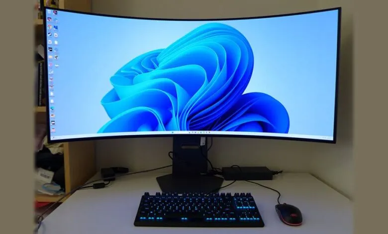 Curved ultrawide monitor displaying Windows 11 wallpaper with a backlit keyboard and mouse.