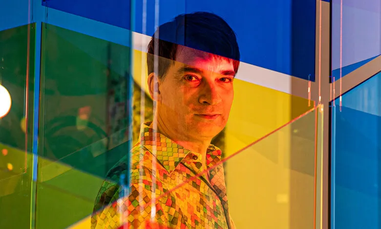 Man with dark hair and a colorful patterned shirt looks towards the camera, framed by abstract colored glass panels.