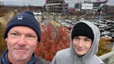 Two people pose in front of Nissan Stadium, Nashville, on an overcast day.