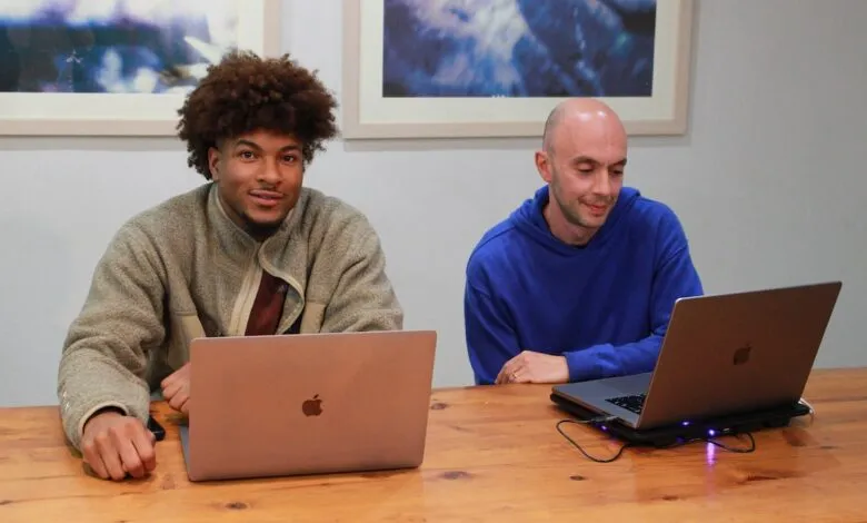 Two men working on laptops at a wooden table, one looking at the camera.