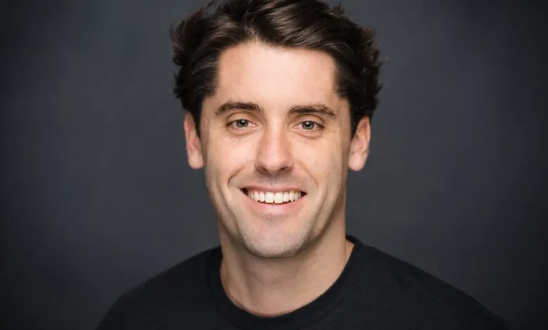 Close-up portrait of a smiling man with dark hair and green eyes against a dark gray background.