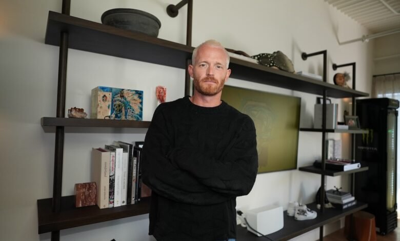Man with blonde hair and beard stands with arms crossed in modern room.