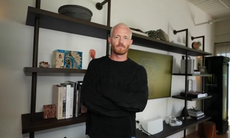 Man with blonde hair and beard stands with arms crossed in modern room.