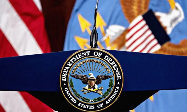 Department of Defense seal on a podium with US flag and military branch flags in the background.