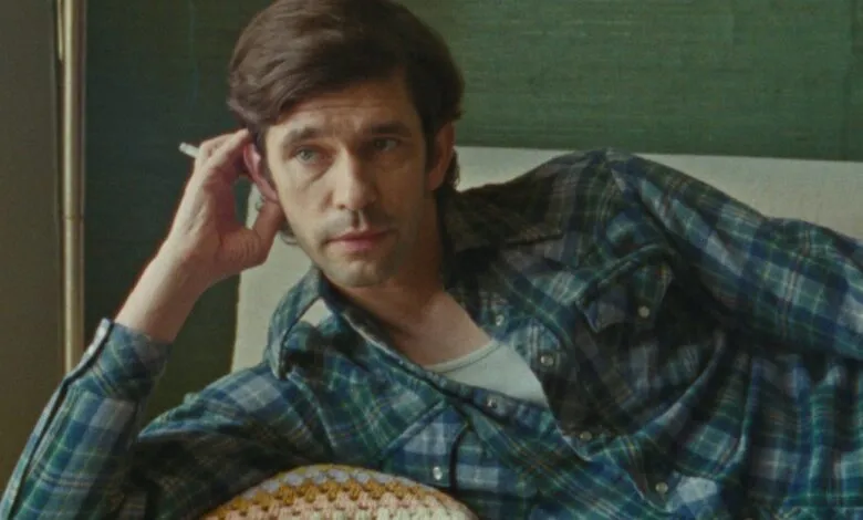 Man with blue eyes and dark hair wearing a plaid shirt, holding a cigarette.