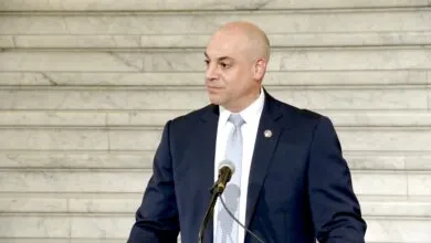 Bald man in a dark suit and light tie speaks at a podium with a microphone.
