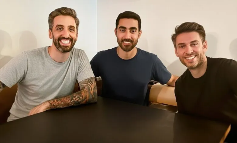 Three smiling men with beards and casual t-shirts sitting at a dark table.