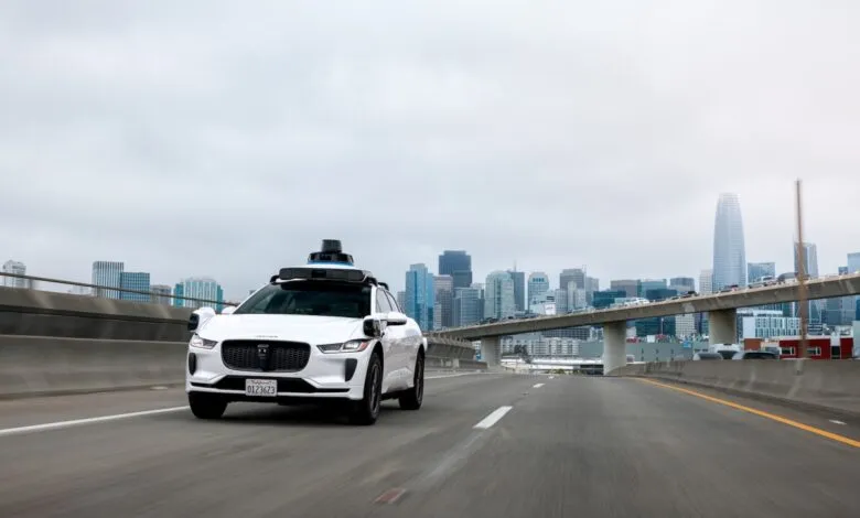 White Waymo self-driving Jaguar I-Pace on freeway with San Francisco skyline.