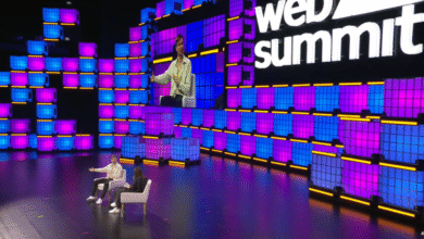 Two people sit on chairs on a stage with a large screen displaying a speaker, illuminated by blue and pink lights.
