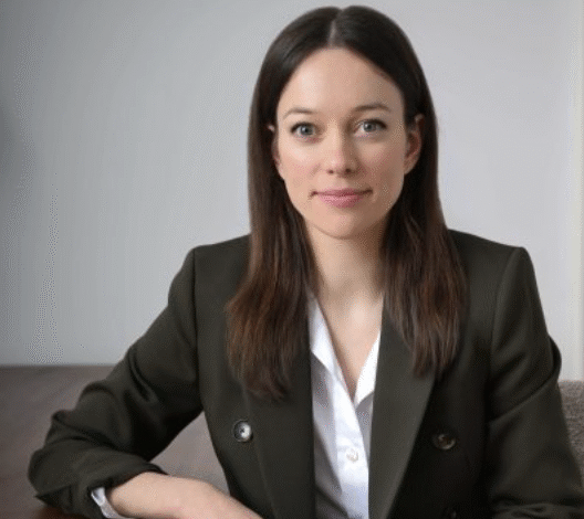 Professional portrait of a woman in a dark green blazer and white shirt, seated at a wooden table.