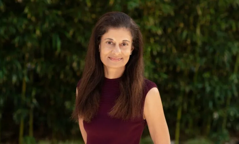Portrait of a woman with long, dark hair smiling outdoors with a blurred green foliage background.