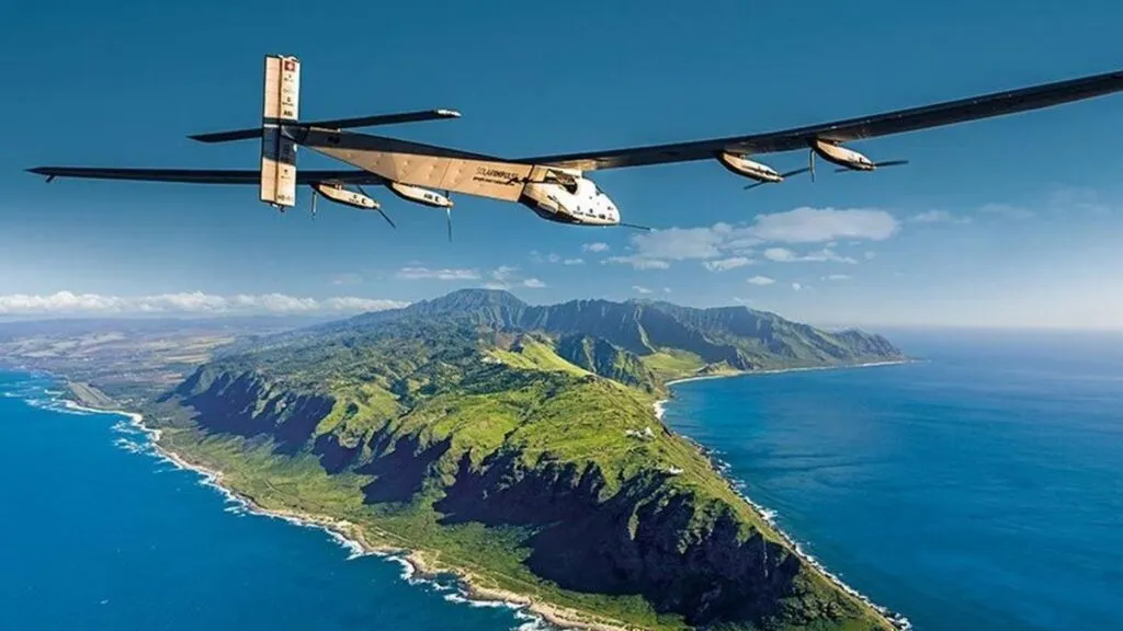 Solar Impulse 2 airplane flying over a lush green coastline with mountains.