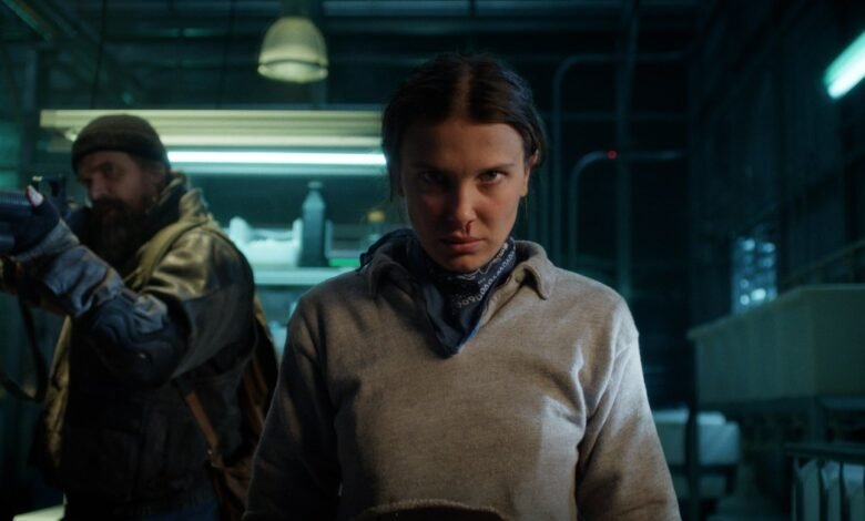 Millie Bobby Brown stares intensely with a man aiming a gun behind her.