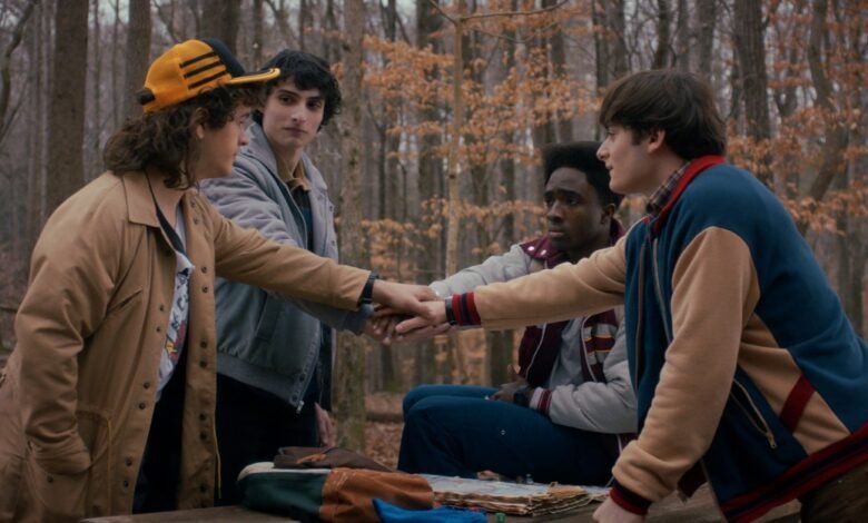 Four young men from Stranger Things stack hands in a forest setting.
