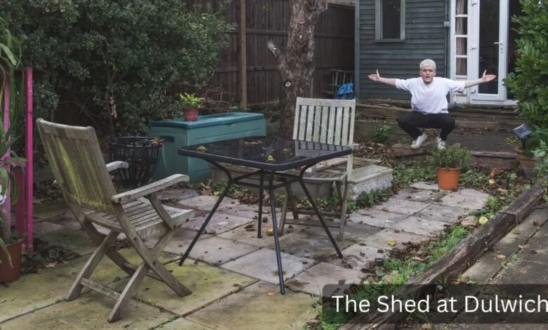 Person squats in garden with arms outstretched near shed, table, and chairs.