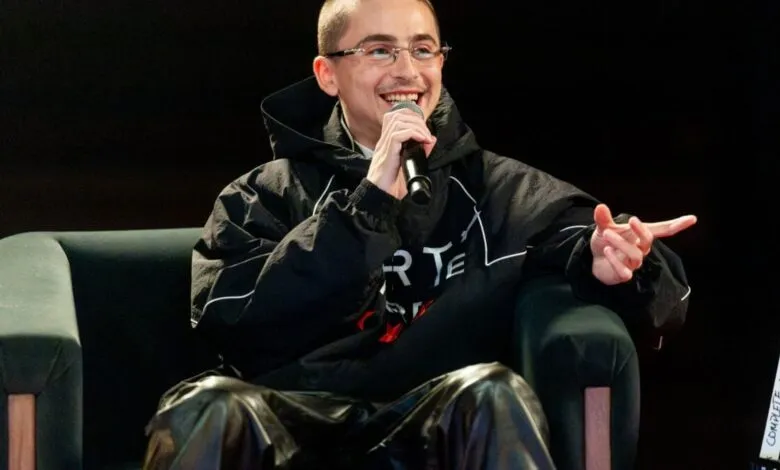 A man with glasses and a microphone smiles while sitting on a dark green couch.