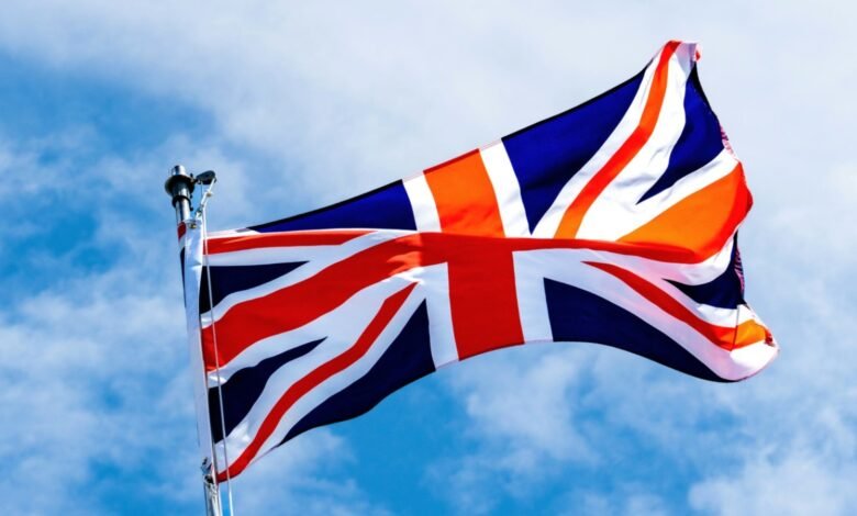 The Union Jack flag of the United Kingdom waving against a bright blue sky with scattered clouds.