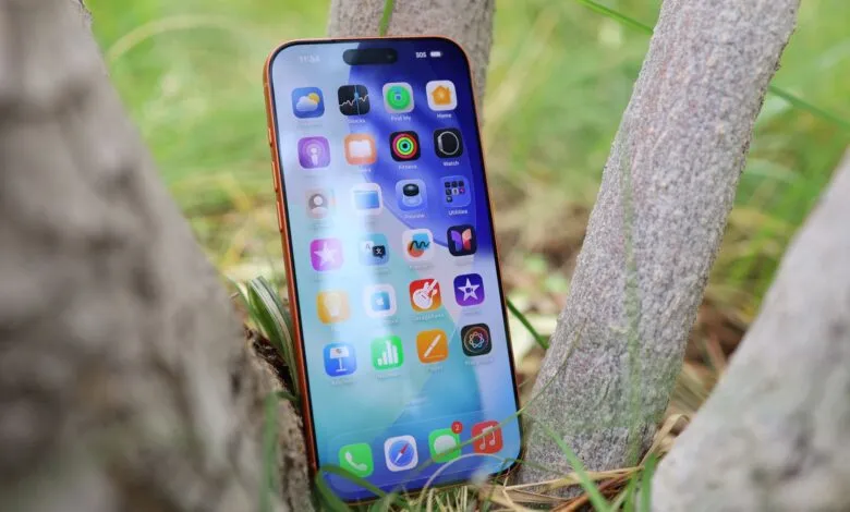 Orange iPhone displaying iOS home screen nestled between tree branches and grass.