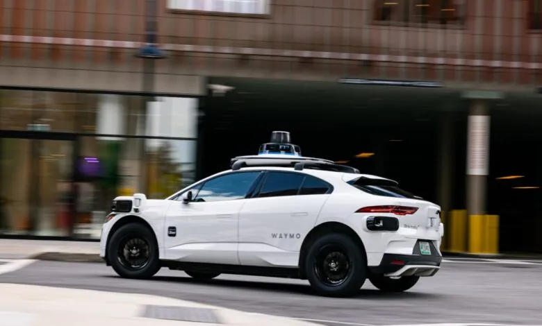 A white Waymo autonomous vehicle drives on a city street with motion blur.