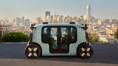 A light teal autonomous Zoox vehicle drives on a street with San Francisco skyline in the background.