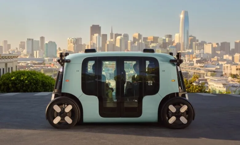 A light teal autonomous Zoox vehicle drives on a street with San Francisco skyline in the background.