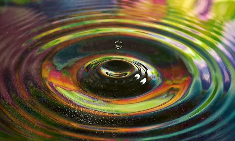 A single water droplet suspended above colorful ripples on a liquid surface.