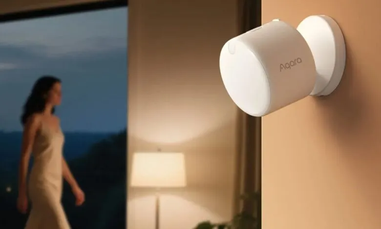 White Aqara motion sensor mounted on a tan wall, with a blurred woman walking in the background.