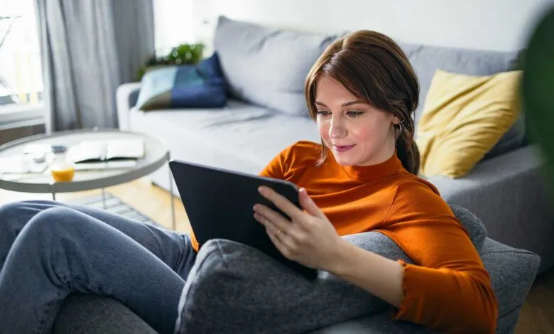 Woman in orange turtleneck using a tablet while relaxing on a sofa.