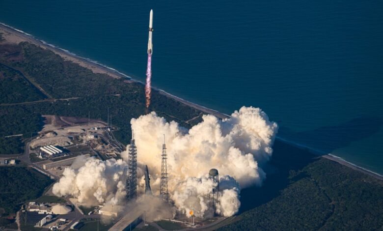 A rocket launches from a coastal launchpad, creating a massive plume of white smoke.