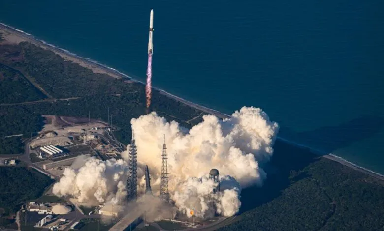 A rocket launches from a coastal launchpad, creating a massive plume of white smoke.