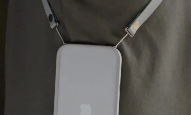 Close-up of a white iPhone with a crossbody strap attached, worn over a dark green shirt.