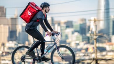 Delivery person on a bicycle with a red insulated bag on their back, city skyline in background.