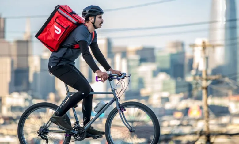 Delivery person on a bicycle with a red insulated bag on their back, city skyline in background.
