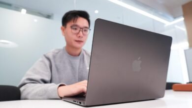 Young man wearing glasses and a grey sweatshirt works on a laptop.